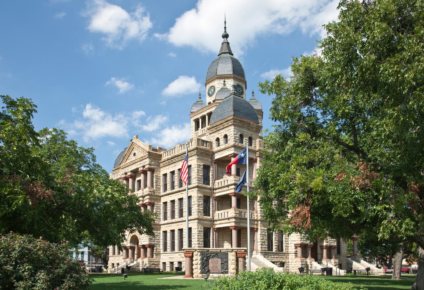 The historic Denton County Courthouse, a stone building with a domed clock tower, framed by mature green trees.