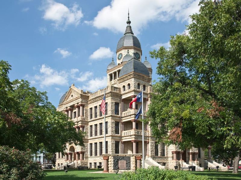 The historic Denton County Courthouse, a stone building with a domed clock tower, framed by mature green trees on the town square.