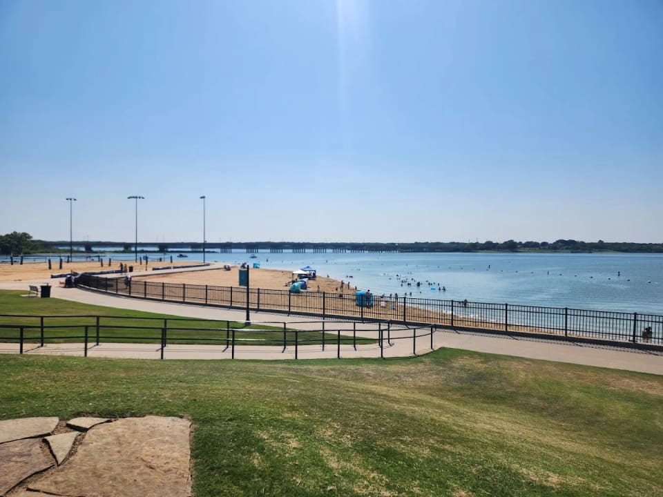 The sandy swimming beach and shoreline of Little Elm Park on Lewisville Lake, with a bridge in the distance under a clear blue sky.